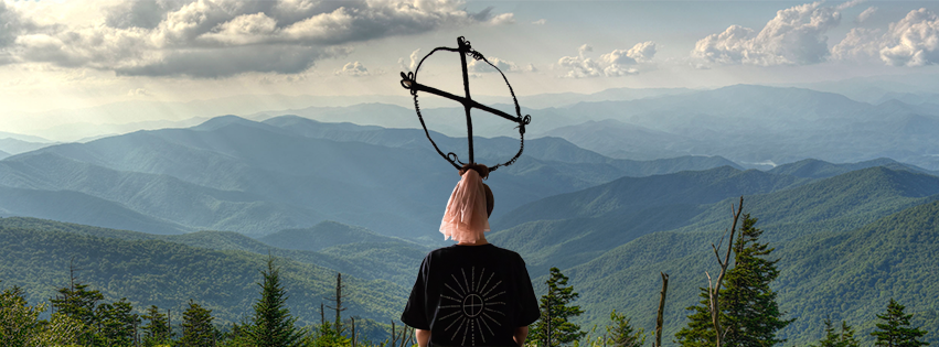 A figure in white facing a mountain range, a sculptural wire form held above their head