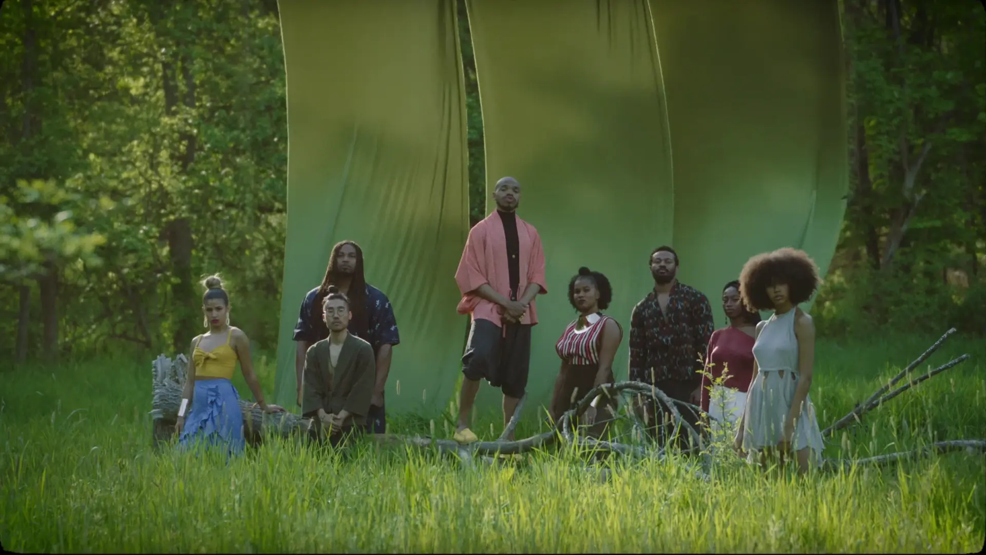 A still from the official music video for bamboo: the full cast gathered among tall green fabric panels in a sunlit field